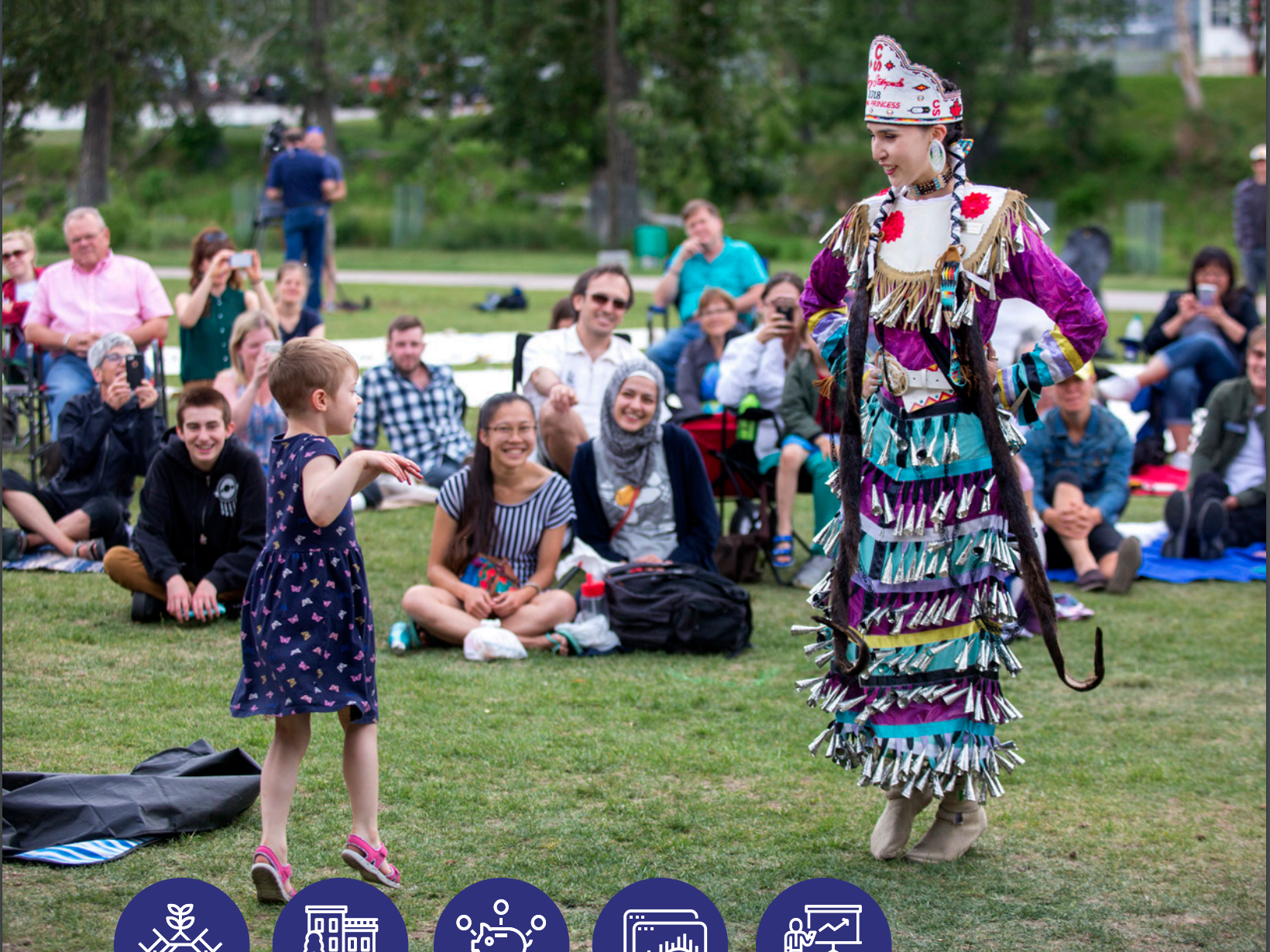 Woman in Indigenous regalia dancing with young caucasian girl in a park while others look on.